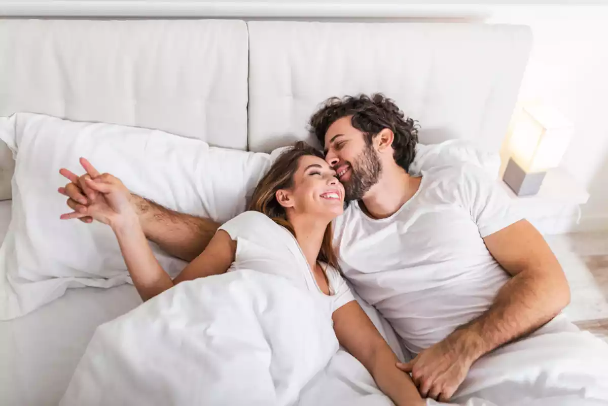 A woman and a man in white clothes hugging each other on a white bed smiling.